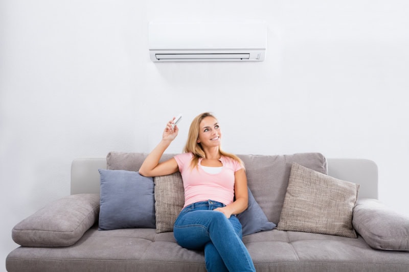Ductless ACs Improve Indoor Air Quality and Control Humidity. Young Happy Woman Sitting On Couch Operating Air Conditioner With Remote Control At Home.