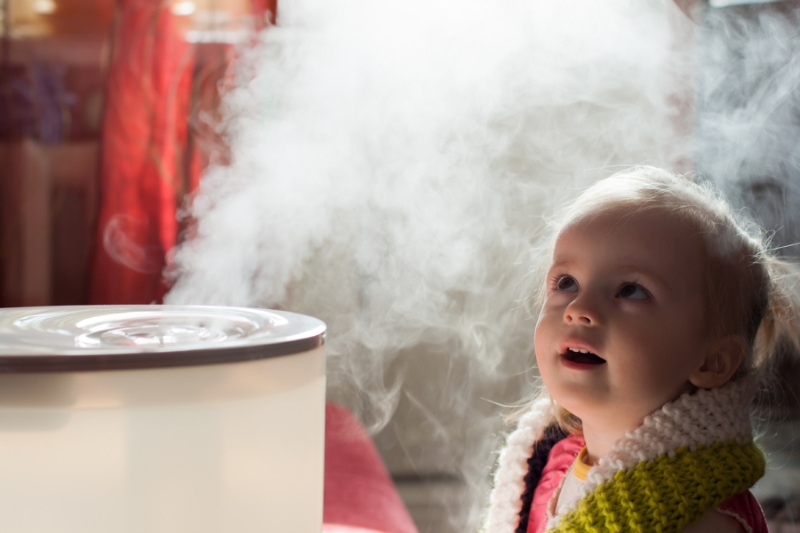 Benefits of Using a Humidifier in Your Home. Image is a photograph of a little girl looking mystified by a humidifier.